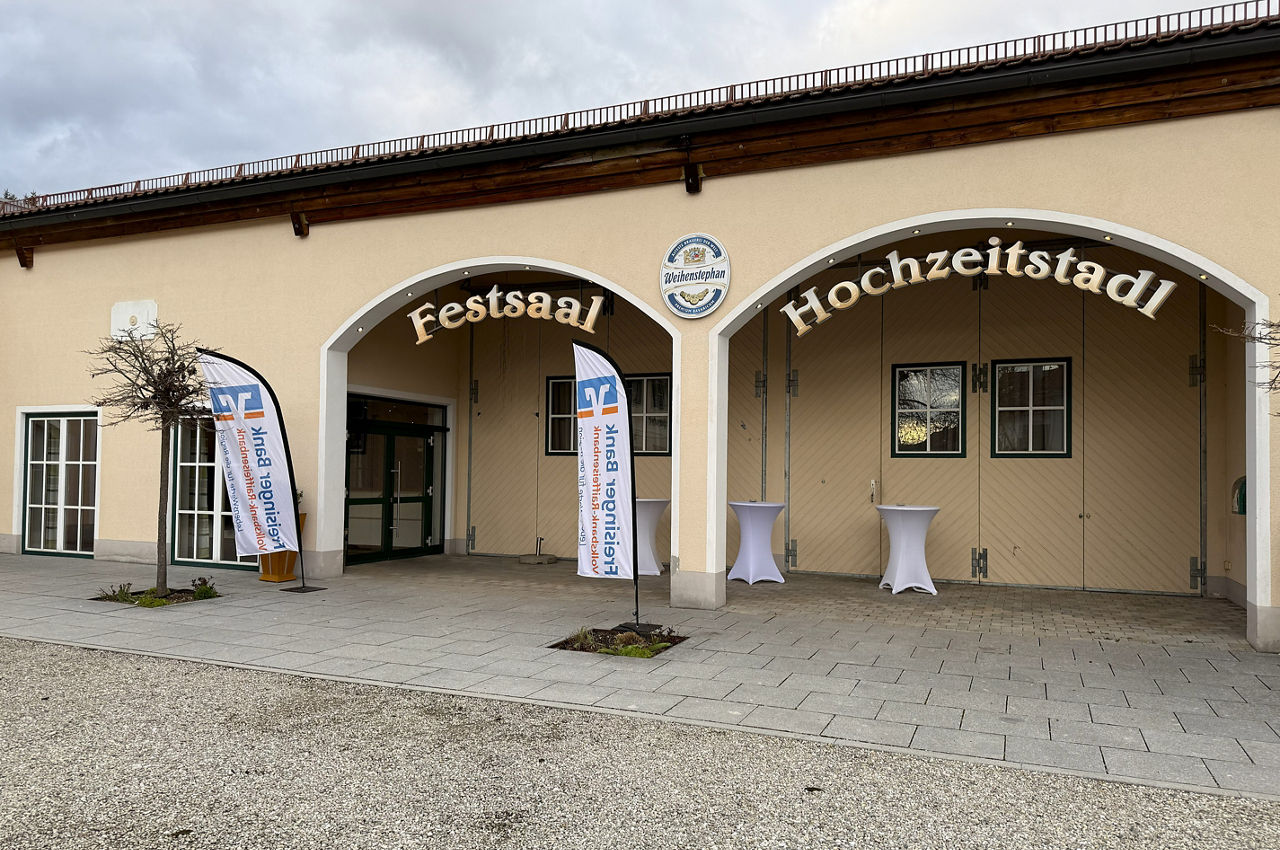 Flaches beiges Gebäude vom Burgerwirt mit Eingang zum Festsaal / Hochzeitsstadl. Davor stehen zwei Beachflags mit dem Logo der Freisinger Bank