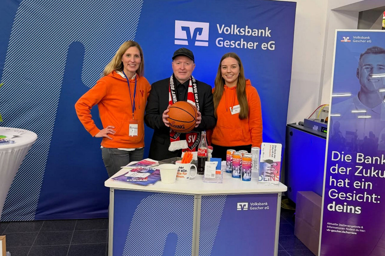 Ein Mann mit Basketball in der Hand steht mit zwei Frauen in orangefarbenen Pullovern vor einer Wand der Volksbank Gescher eG