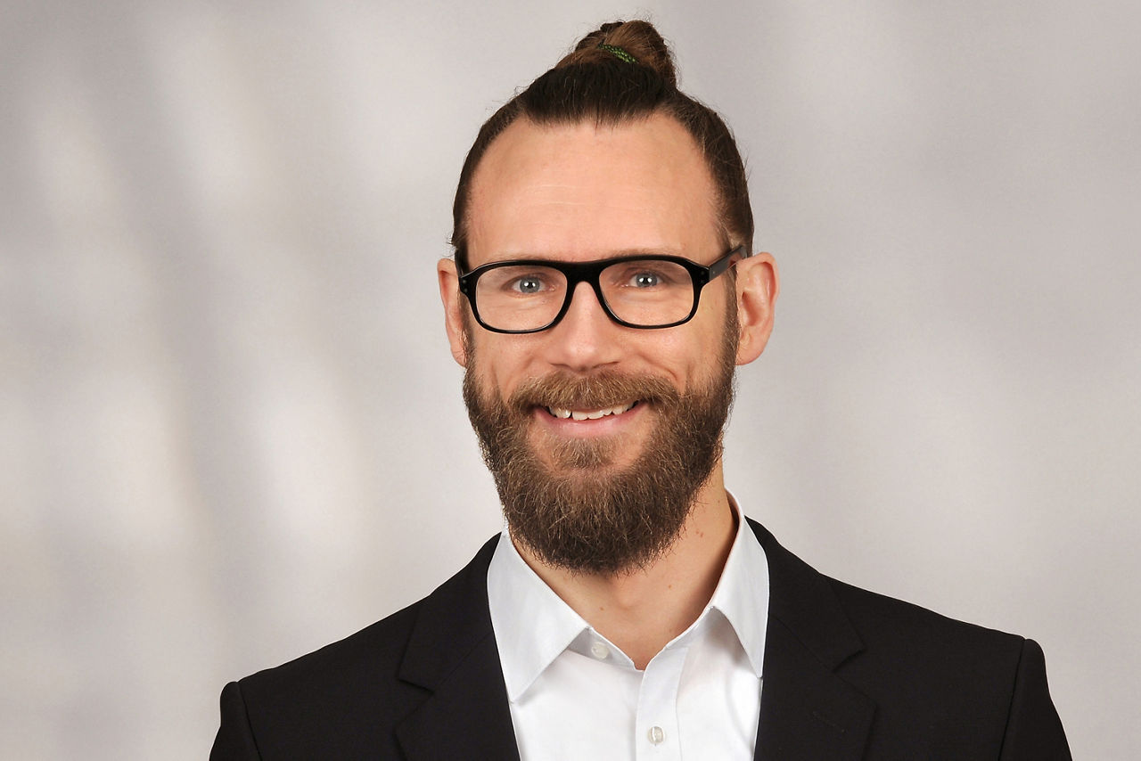 Sebastian Buyting hat braune Haare und diese zu einem Dutt gebunden. Er trägt eine schwarze Brille und einen Bart sowie einen Anzug