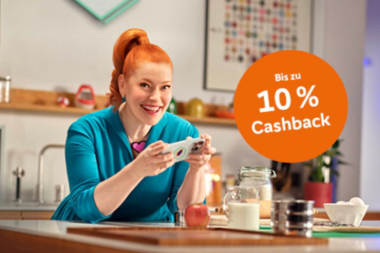 Enie van de Meiklokjes sitzt mit einem Handy in der Hand an einem Küchentisch und lächelt in die Kamera.