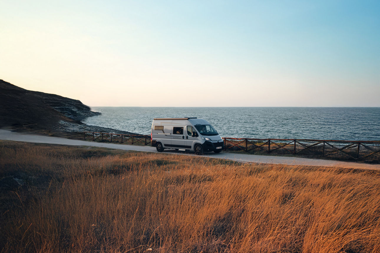 Wohnmobil steht auf einem Weg am Meer mit Gras im Vordergrund und Hügeln im Hintergrund bei klarem Himmel