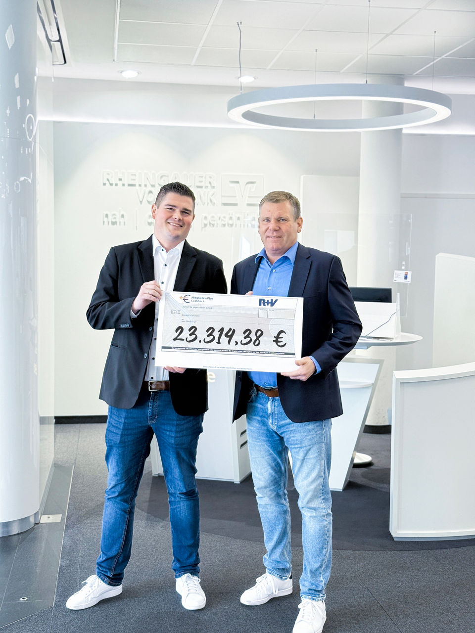 Zwei Männer in Jeans und Sakko halten einen großen Scheck mit der Summe '23.314,38 €' in einem modernen Büro mit weißer Einri