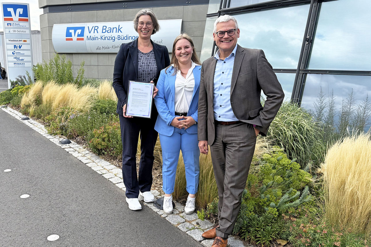 VR Bank Main-Kinzig-Büdingen unterzeichnet „Charta der Vielfalt“ – drei Personen präsentieren die Urkunde vor dem Gebäude.