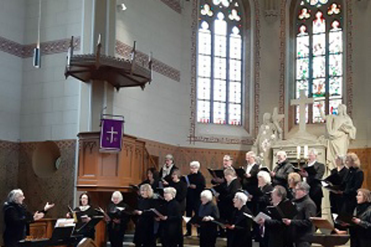 Schwarz gekleidete Choristen stimmen in einer Kirche das Konzert an. Ein Dirigent im Vordergrund sorgt für guten Takt
