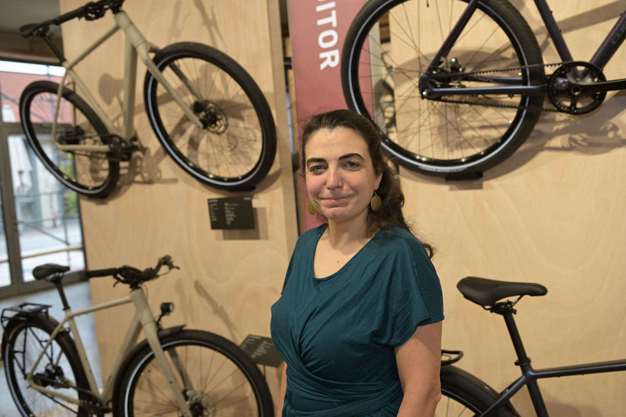 Eine Frau in dunkelgrünem Kleid steht in einem Fahrradgeschäft. Hinter ihr eine Wand mit aufgehängten Rädern