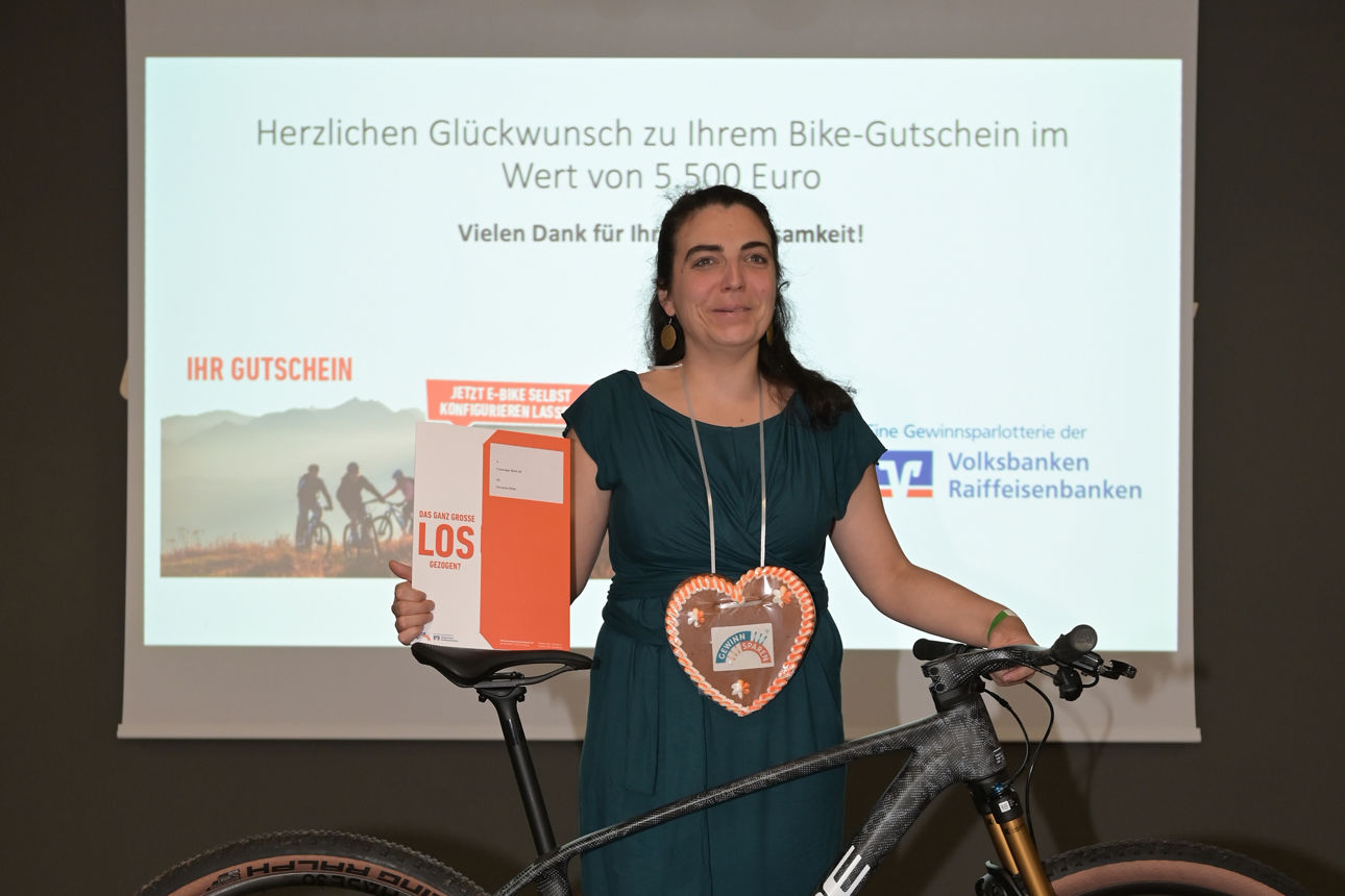 Eine Frau in dunkelgrünem Kleid mit Lebkuchenherz um den Hals und Gutschein in der Hand stehe hinter einem schwarzen Mountainbike vor einer Leinwand mit einem Präsentationsbild