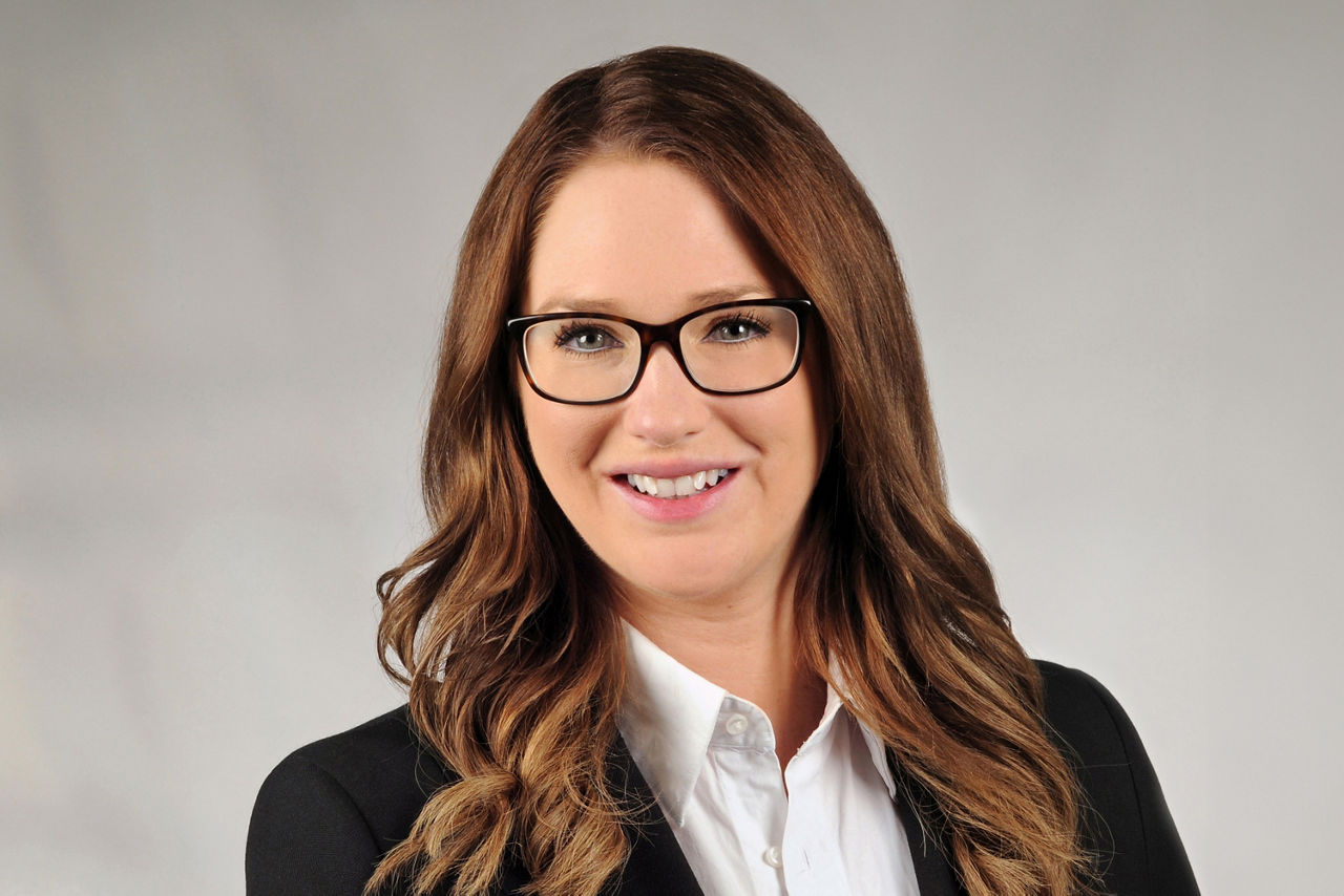 Aileen Clarke hat braune lange leicht gelockte Haare und trägt eine schwarze eckige Brille und ein Businessoutfit 