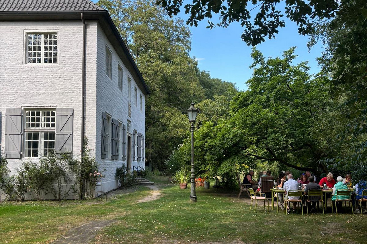 Ein altes weißes Haus. Rechts davon essen Menschen am Tisch.