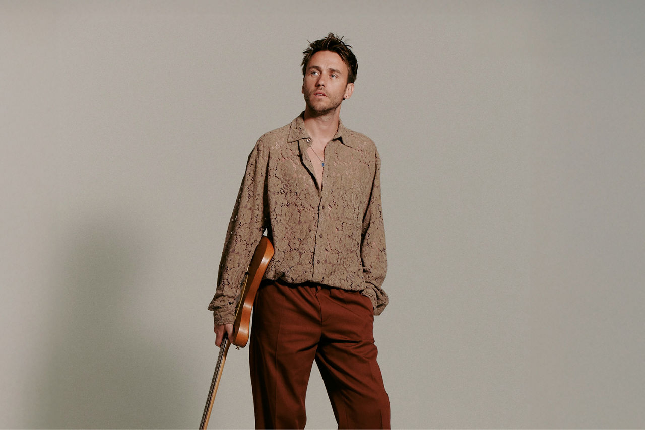 Sänger, Songwriter und Produzent Clueso steht mit einer Gitarre in der Hand vor einem neutralen Hintergrund und schaut ins Nichts.