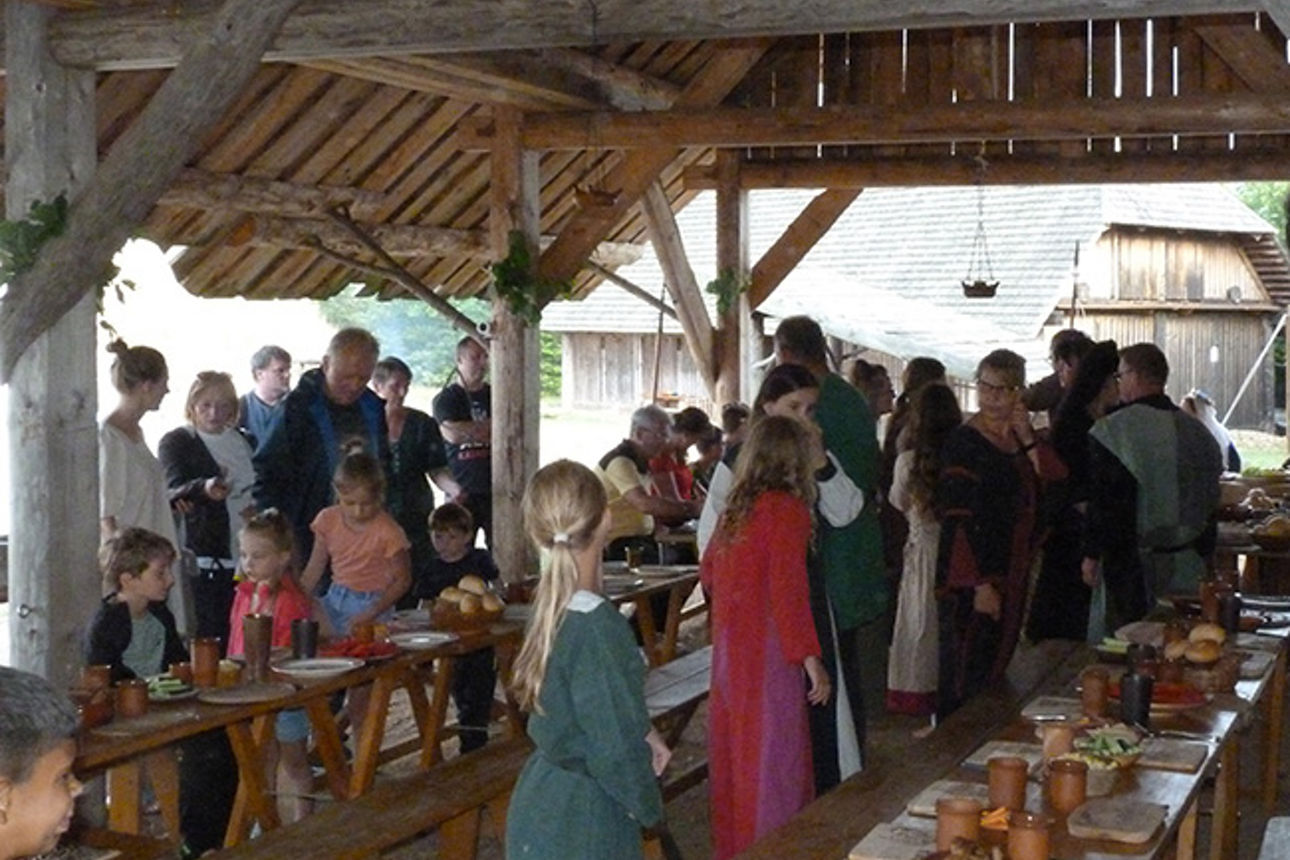 Menschen in mittelalterlicher Kleidung stehen an Tischen in einem Holzpavillon im Freien