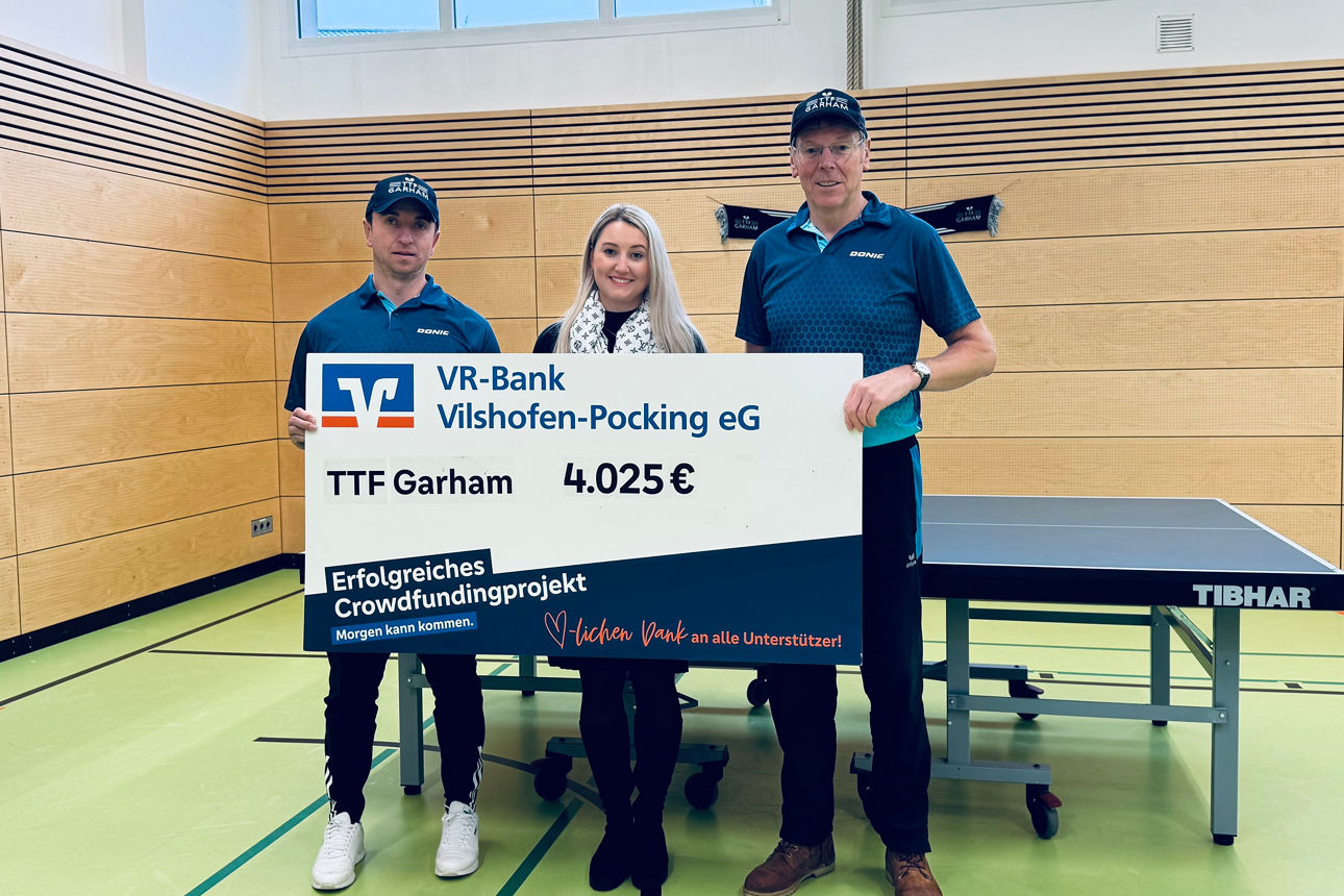 Zwei Männer und eine Frau mit Spendenscheck vor einer Tischtennisplatte