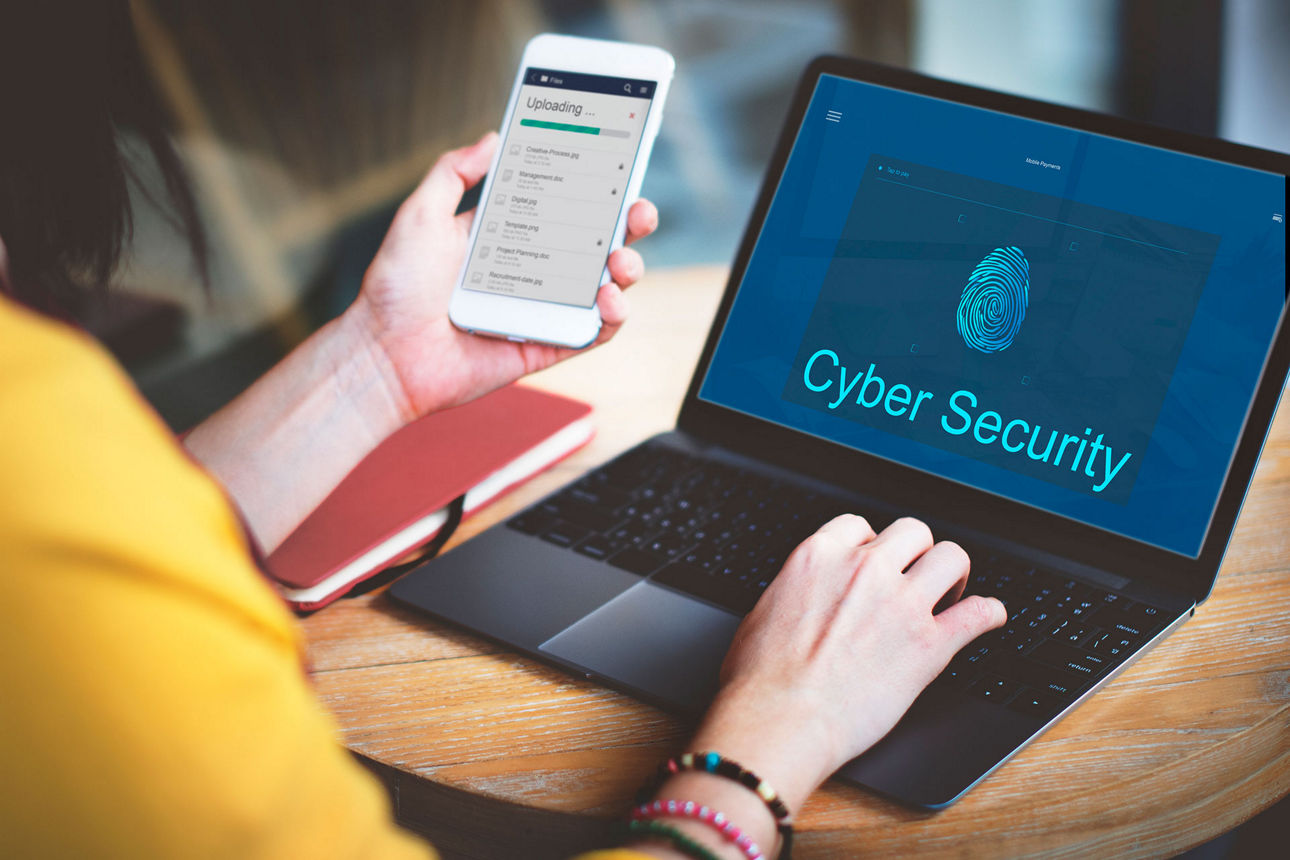 Person hält Smartphone in der einen Hand und die andere auf einem Laptop - Schriftzug "Cyber Security"