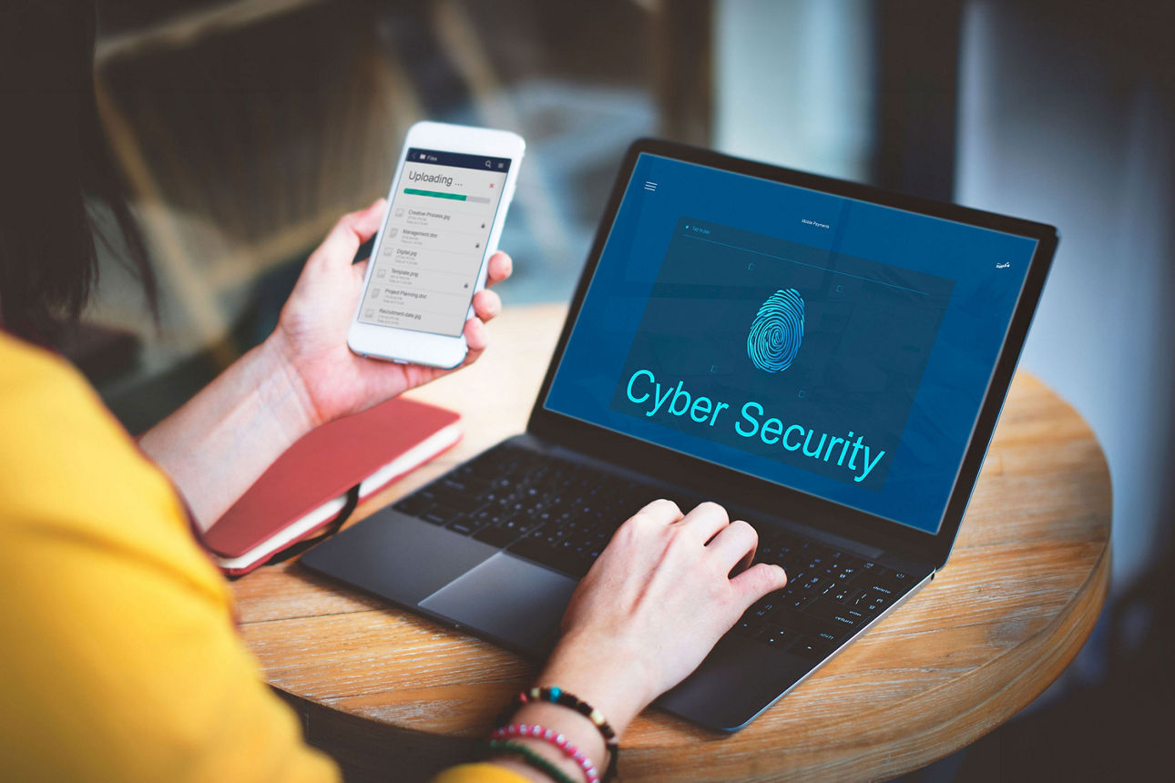 Person hält Smartphone in der einen Hand und die andere auf einem Laptop - Schriftzug "Cyber Security"