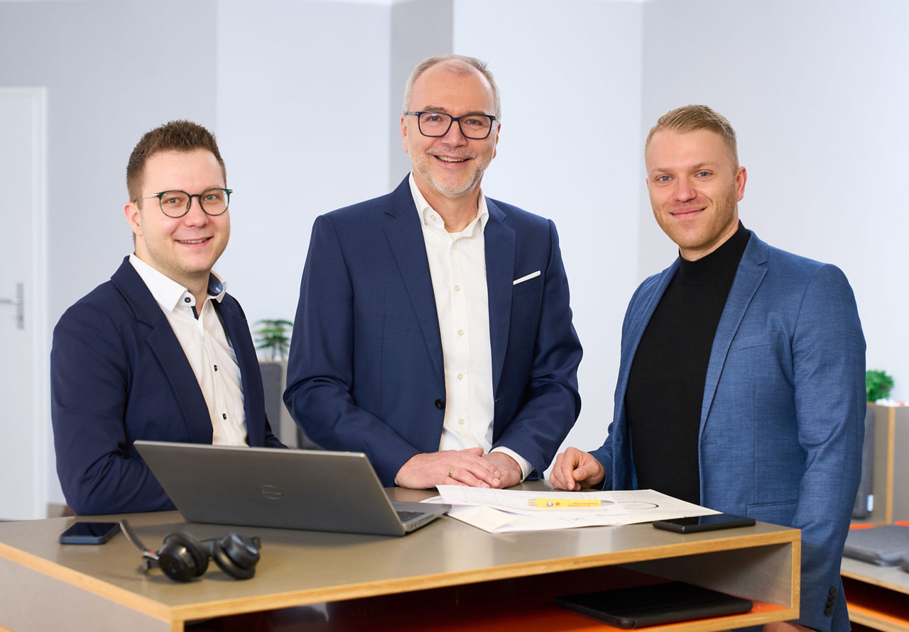 Drei Mitarbeiter der Abteilung RefiLG stehen nebeneinander an einem Tisch mit Laptop und Unterlagen