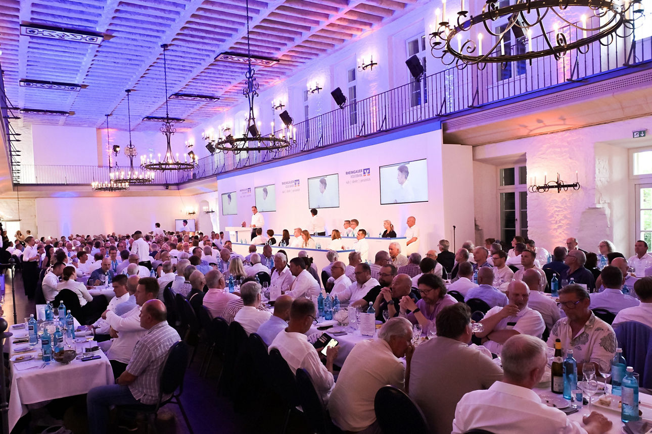 Großer festlich gedeckter Saal mit vielen Personen an langen Tischen und einer Bühne mit mehreren Bildschirmen an der Wand