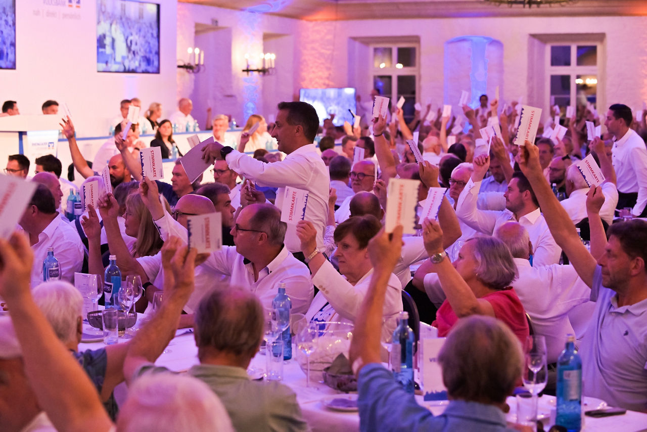 Menschen in einem festlich beleuchteten Saal heben bei der Vertreterversammlung Wahlkarten hoch