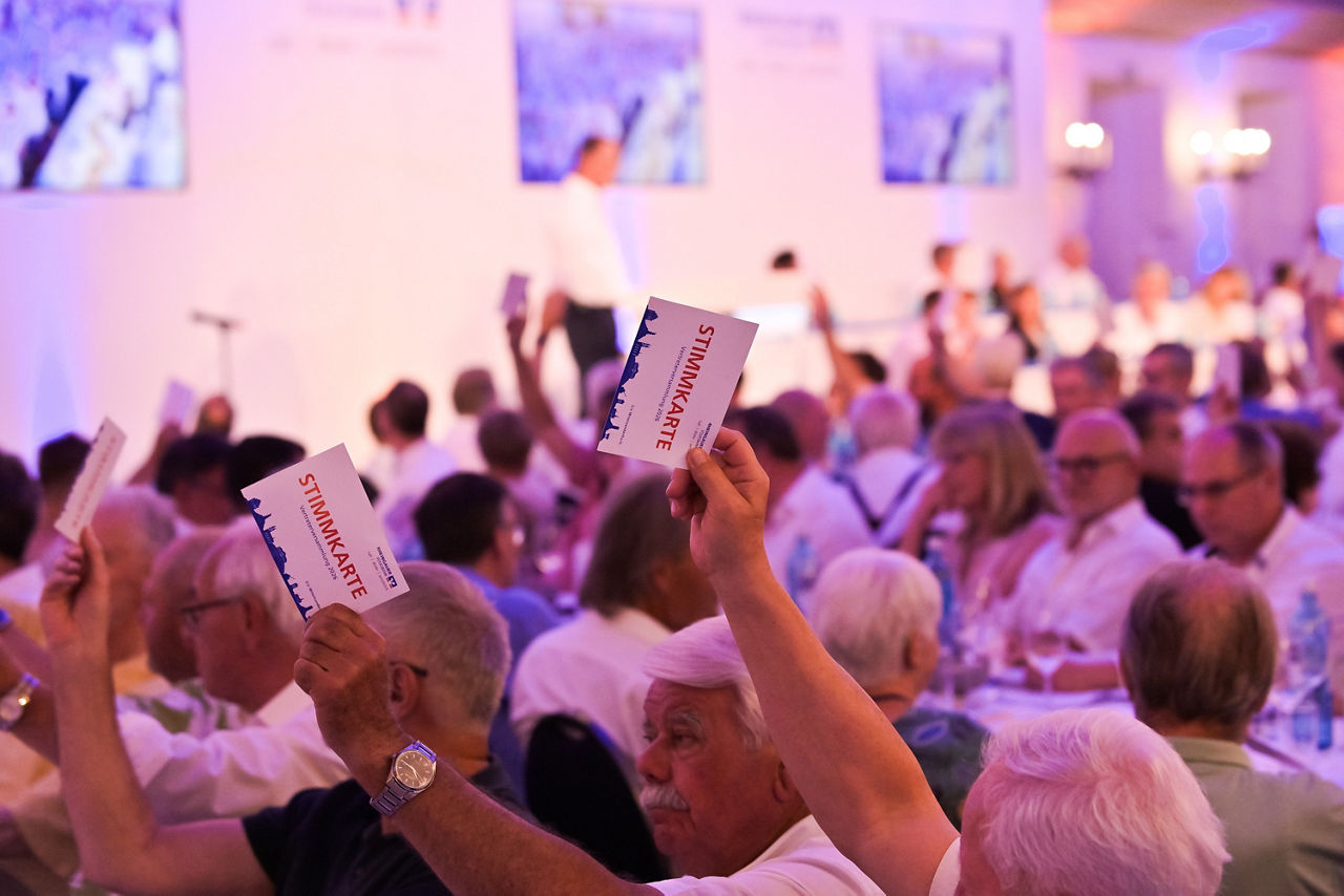Mehrere Personen in einem Saal heben Stimmkarten hoch, im Hintergrund sind weitere Menschen und Bildschirme sichtbar