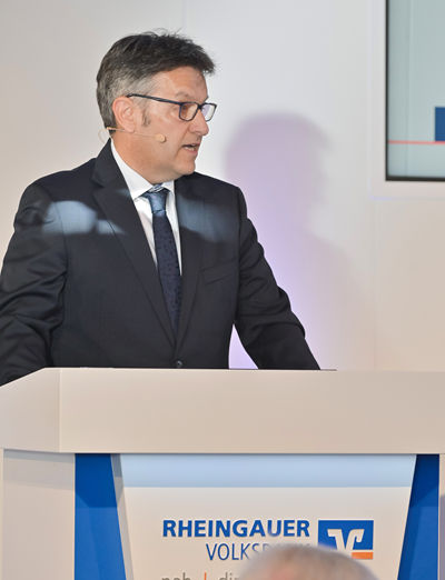 Herr Mager steht am Rednerpult und spricht. Er trägt einen dunklen Anzug mit Krawatte