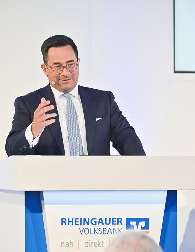Herr Zeiselmaier steht am Rednerpult und spricht. Er trägt einen dunklen Anzug mit weißem Hemd und blauer Krawatte