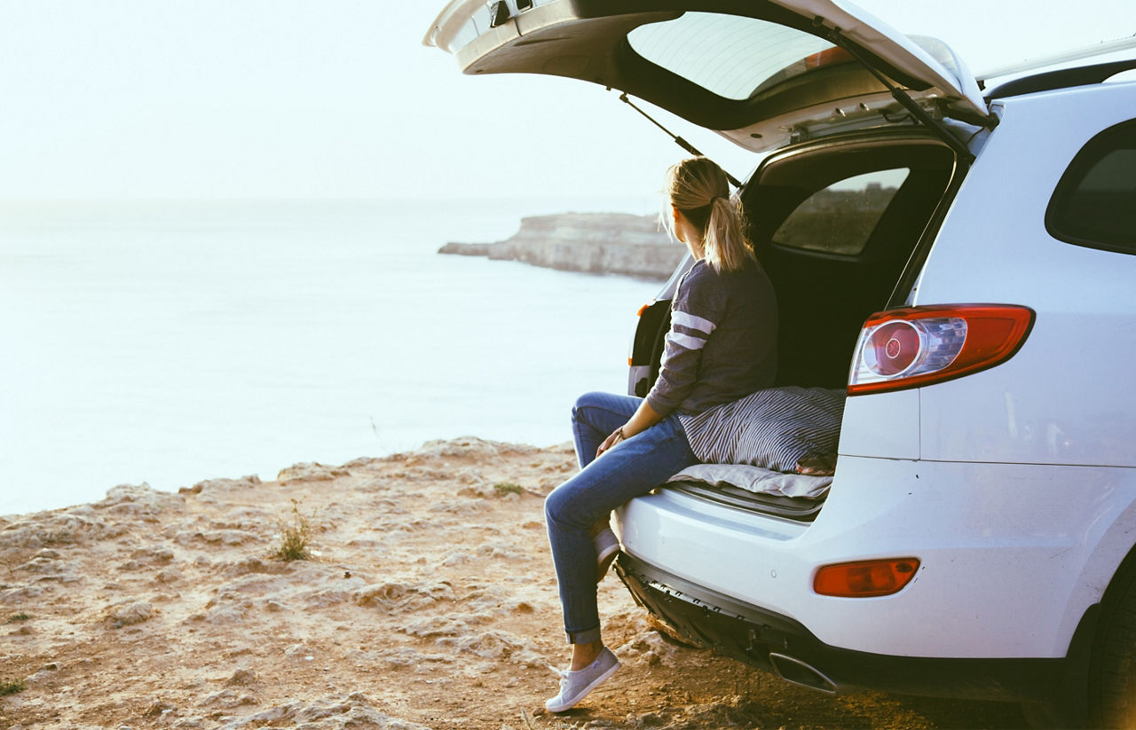 Junge Frau sitzt an einem Kofferraum eines Autos, welches am Meer steht