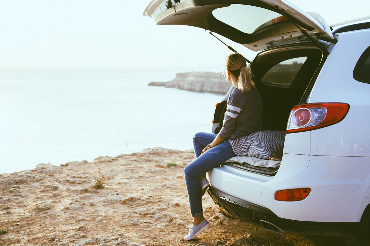 Junge Frau sitzt an einem Kofferraum eines Autos, welches am Meer steht