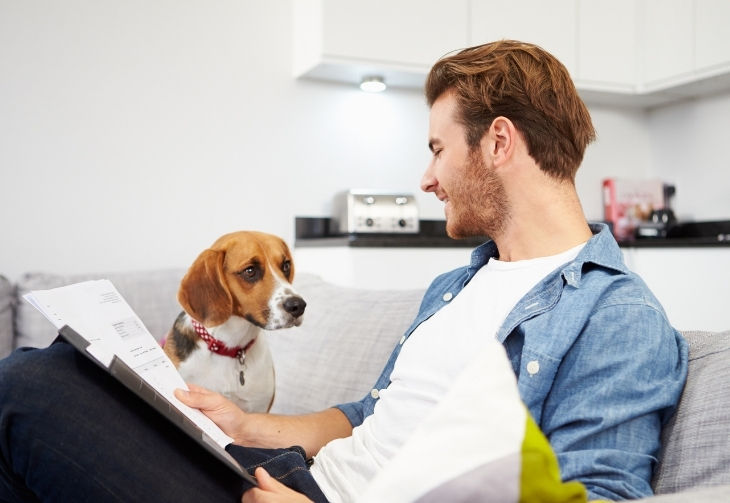 Junger Mann mit Unterlagen in der Hand sitzt mit einem Hund auf einem Sofa