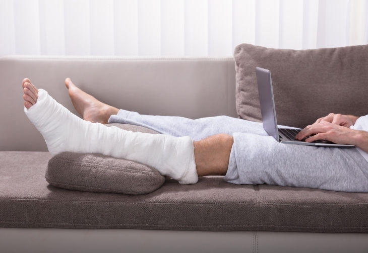Person liegt mit einem Gips am Fuß und einem Laptop auf dem Schoß auf einem Sofa