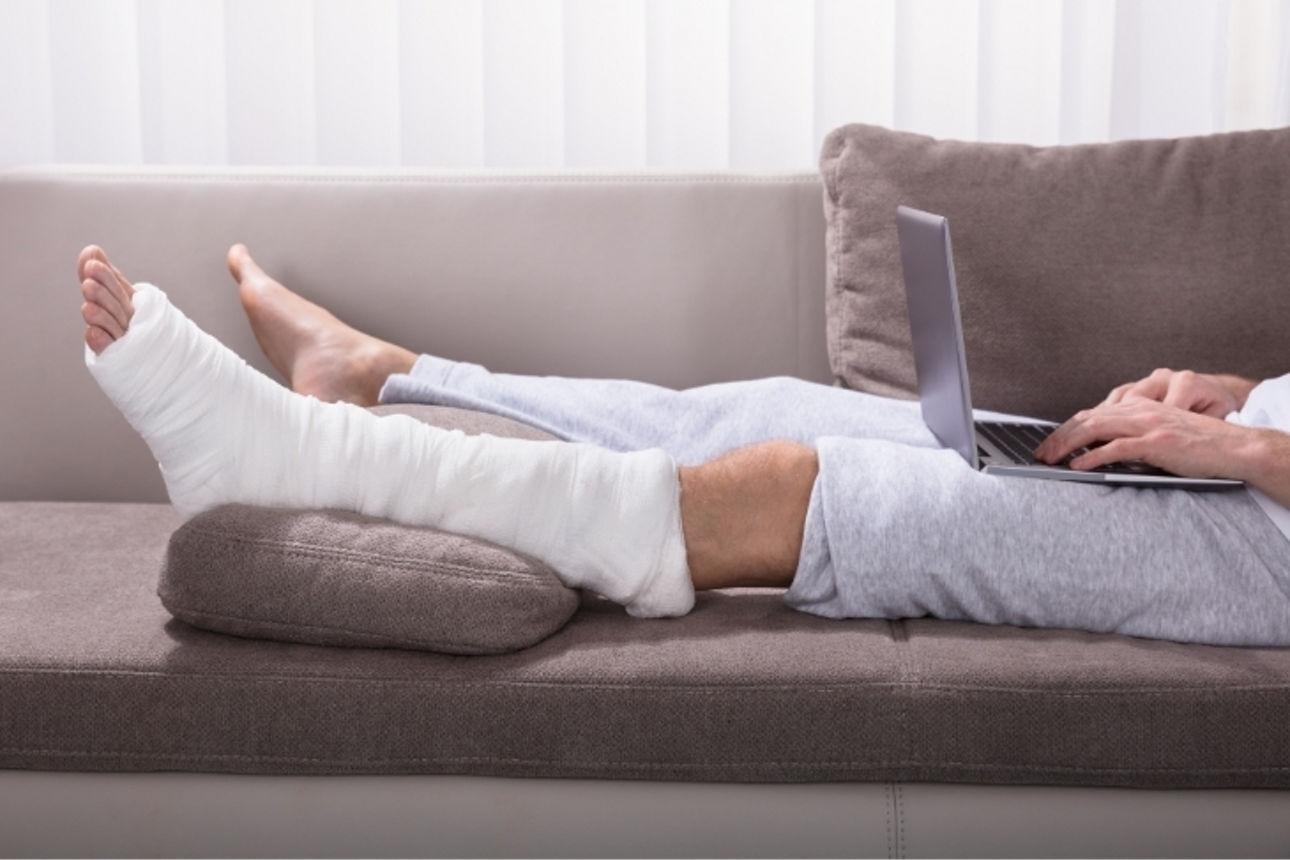 Person liegt mit einem Gips am Fuß und einem Laptop auf dem Schoß auf einem Sofa