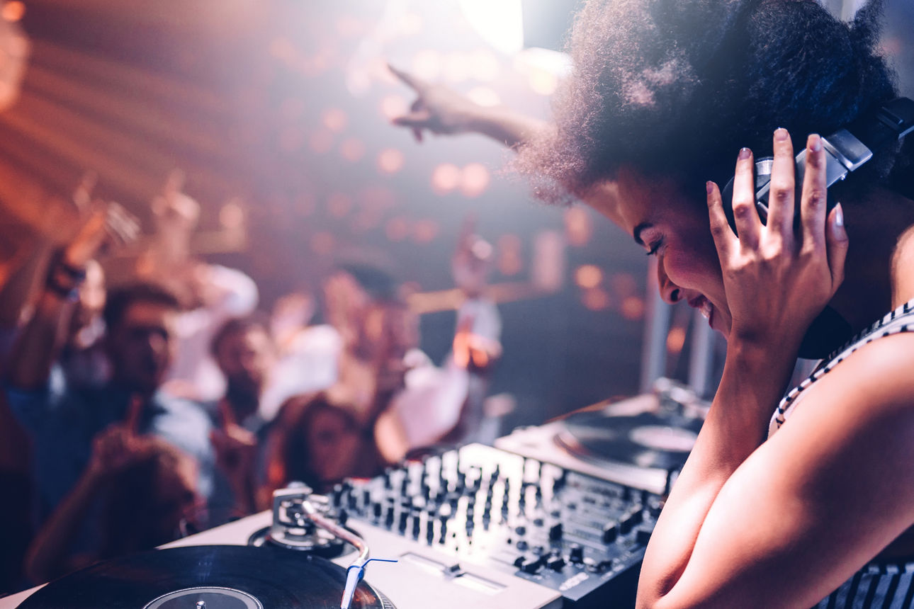 Person bedient Plattenspieler und Mischpult vor Publikum in einem Club mit Bühnenlicht.