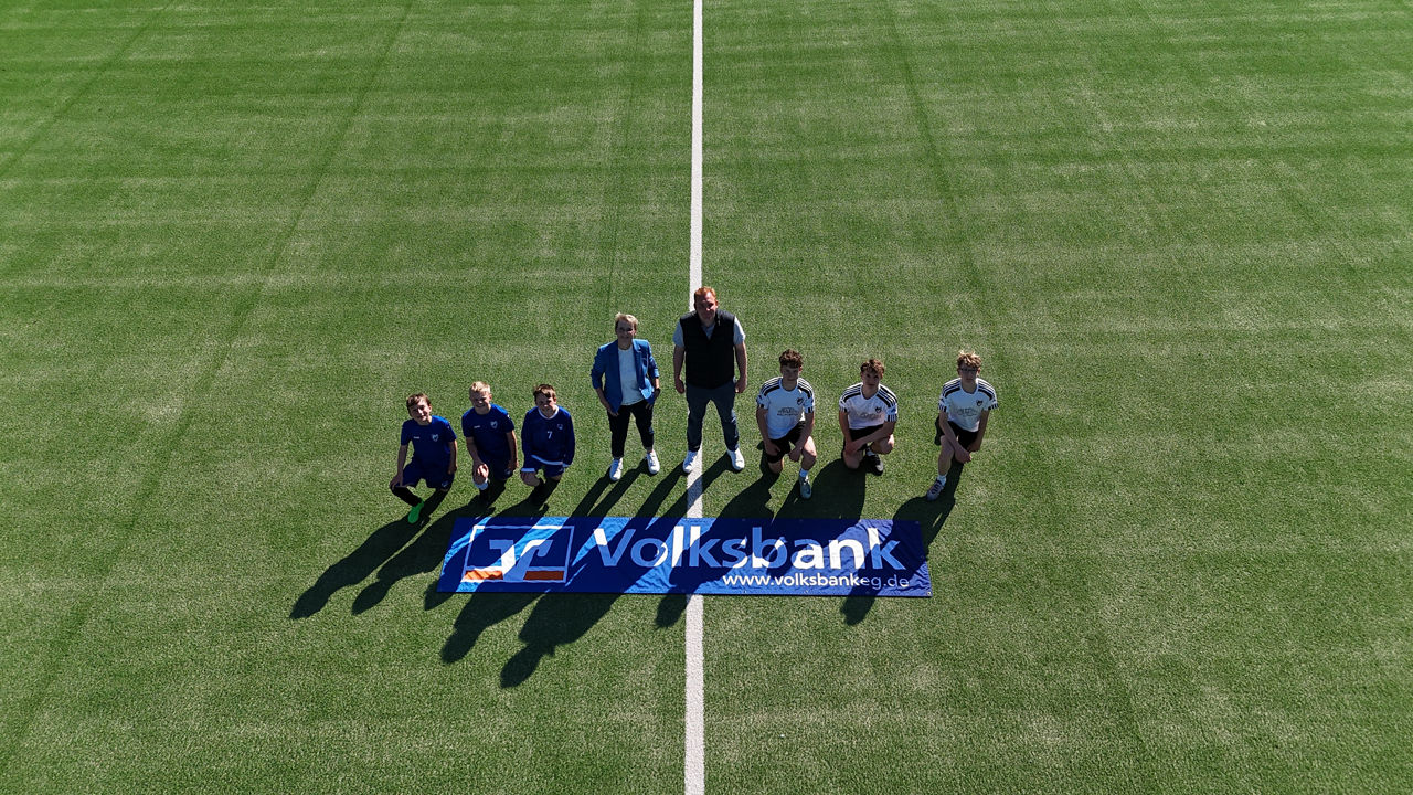 Kinder und ein Mann auf einem Fußballfeld und auf dem Spielfeld liegt eine blaue Volksbank Flagge. 