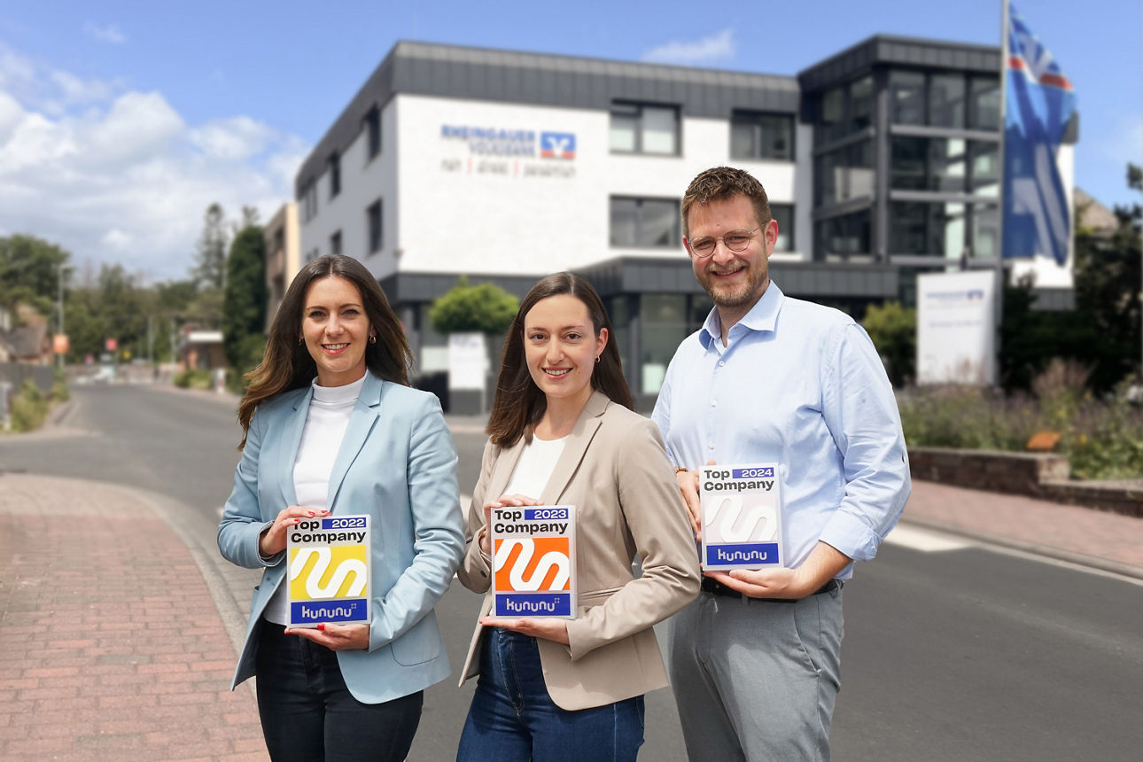 Drei Personen stehen vor der Hauptstelle der Rheingauer Volksbank und halten Auszeichnungen von kununu in den Händen