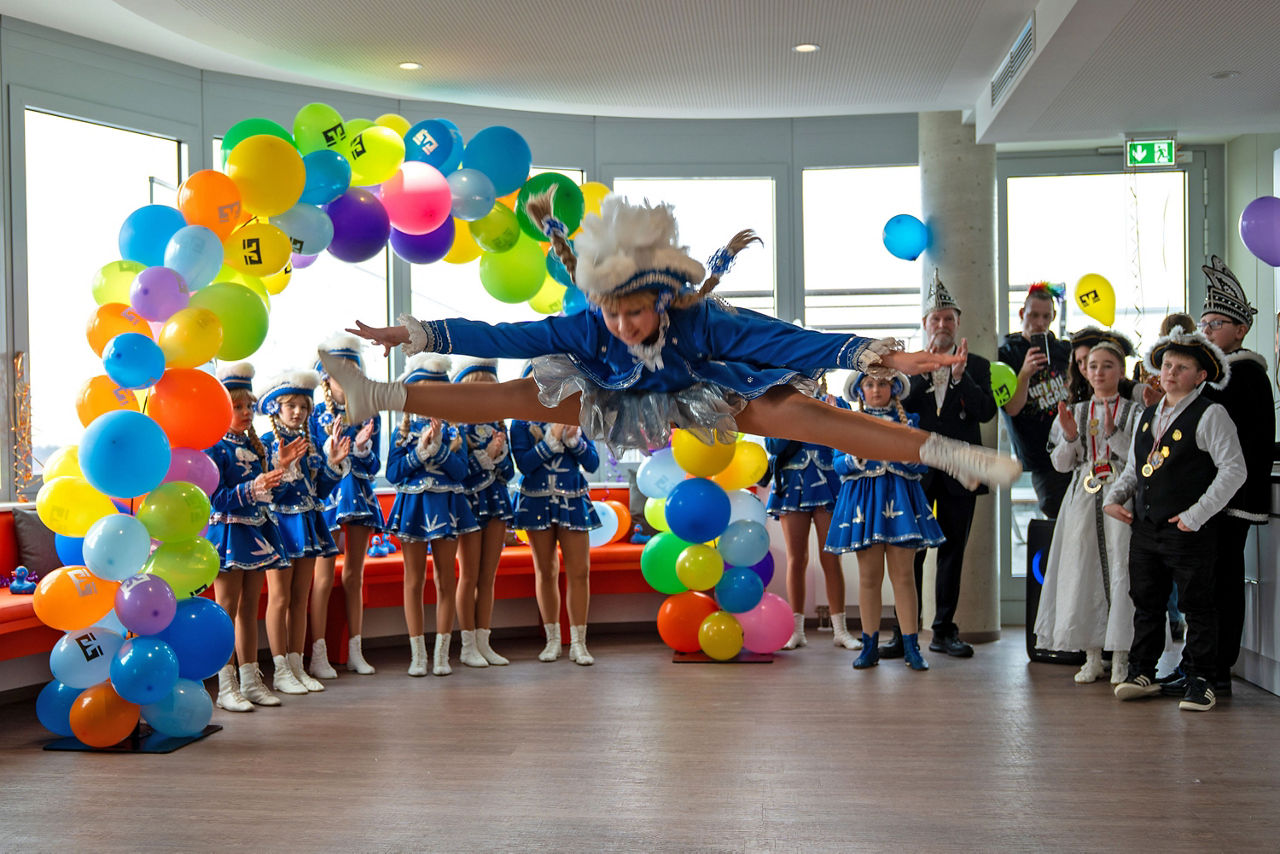 Gardetänzerin im Spagatsprung unter buntem Luftballonbogen, umrahmt von jubelnden Gruppen.