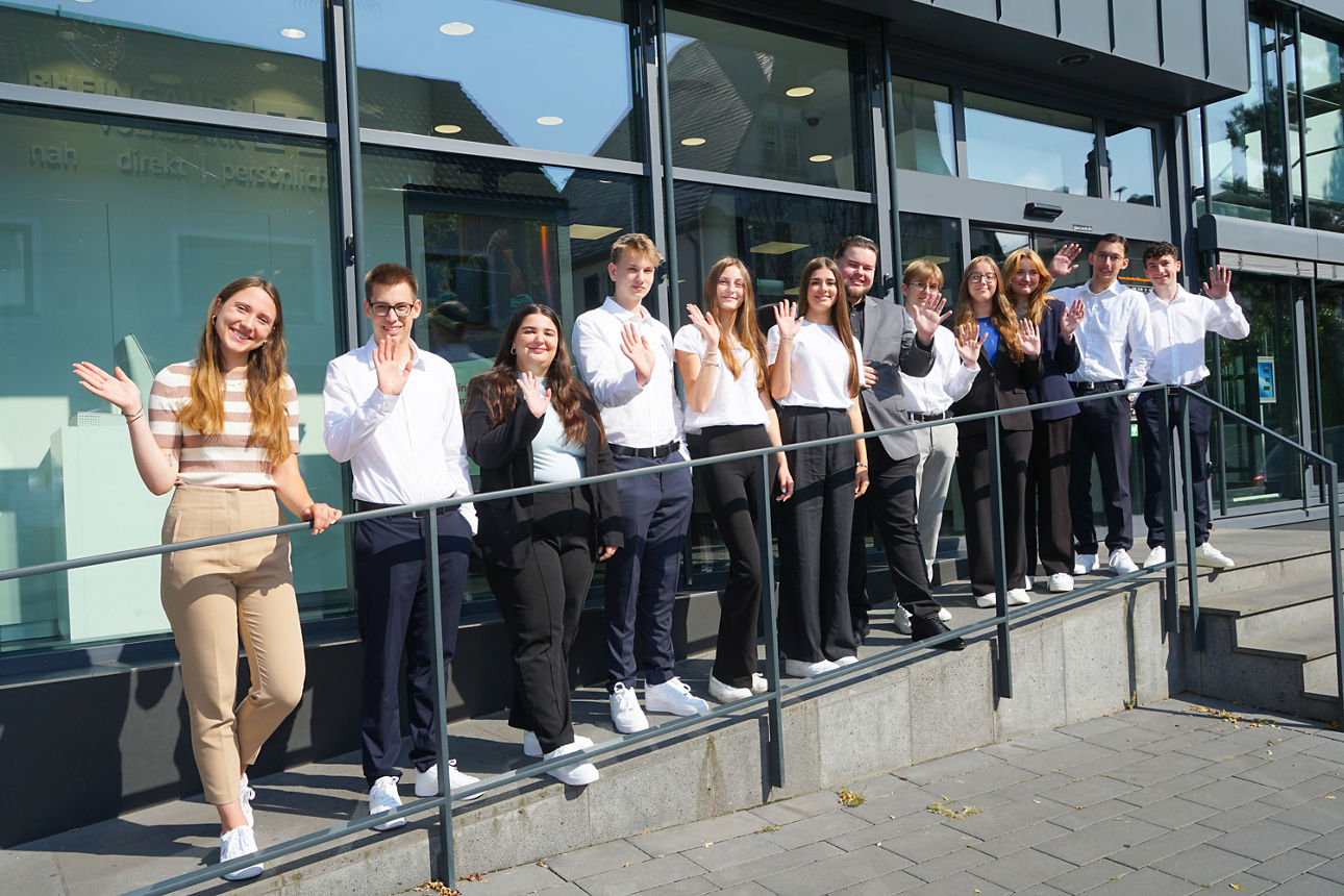 Auszubildende der Rheingauer Volksbank eG stehen vor der Bank in einer Reihe und winken.