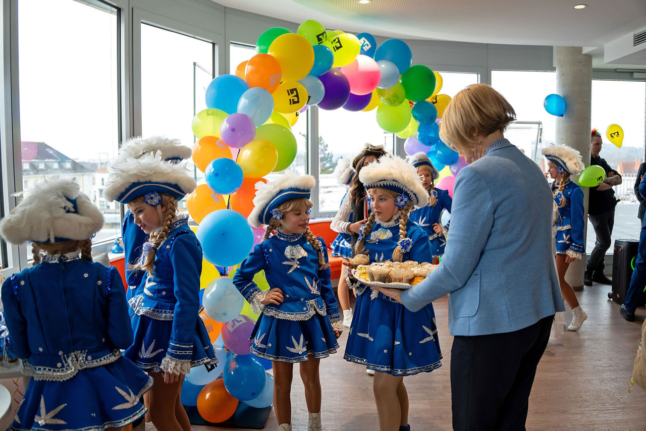 Kinder in blauen Gardekostümen erhalten Gebäck unter buntem Luftballonbogen.