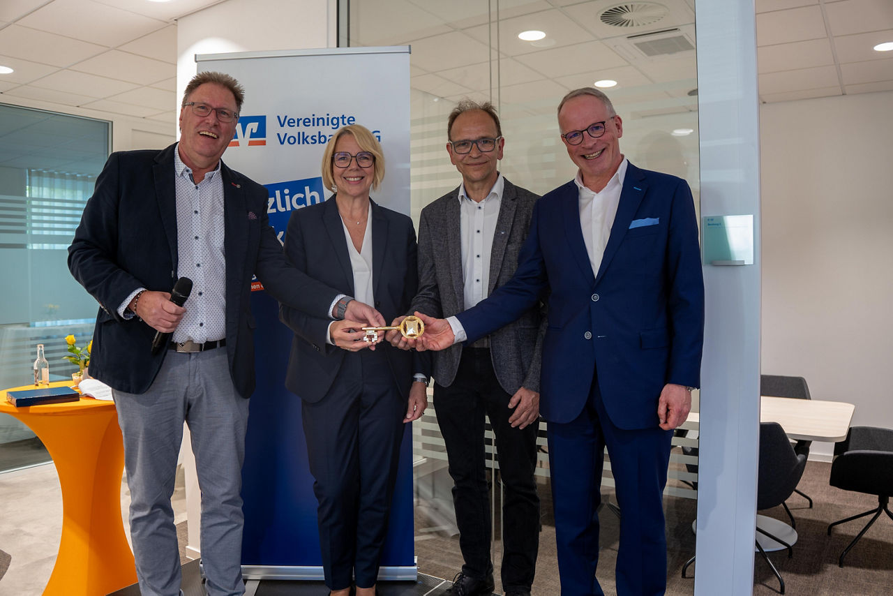 Vier Personen lächeln vor einem Roll-up der „Vereinigte Volksbanken“; sie halten gemeinsam zwei kleine, runde Abzeichen in di