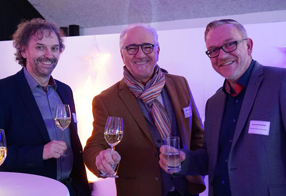 Drei Männer lachen in die Kamera und halten ein Glas Wein in der Hand