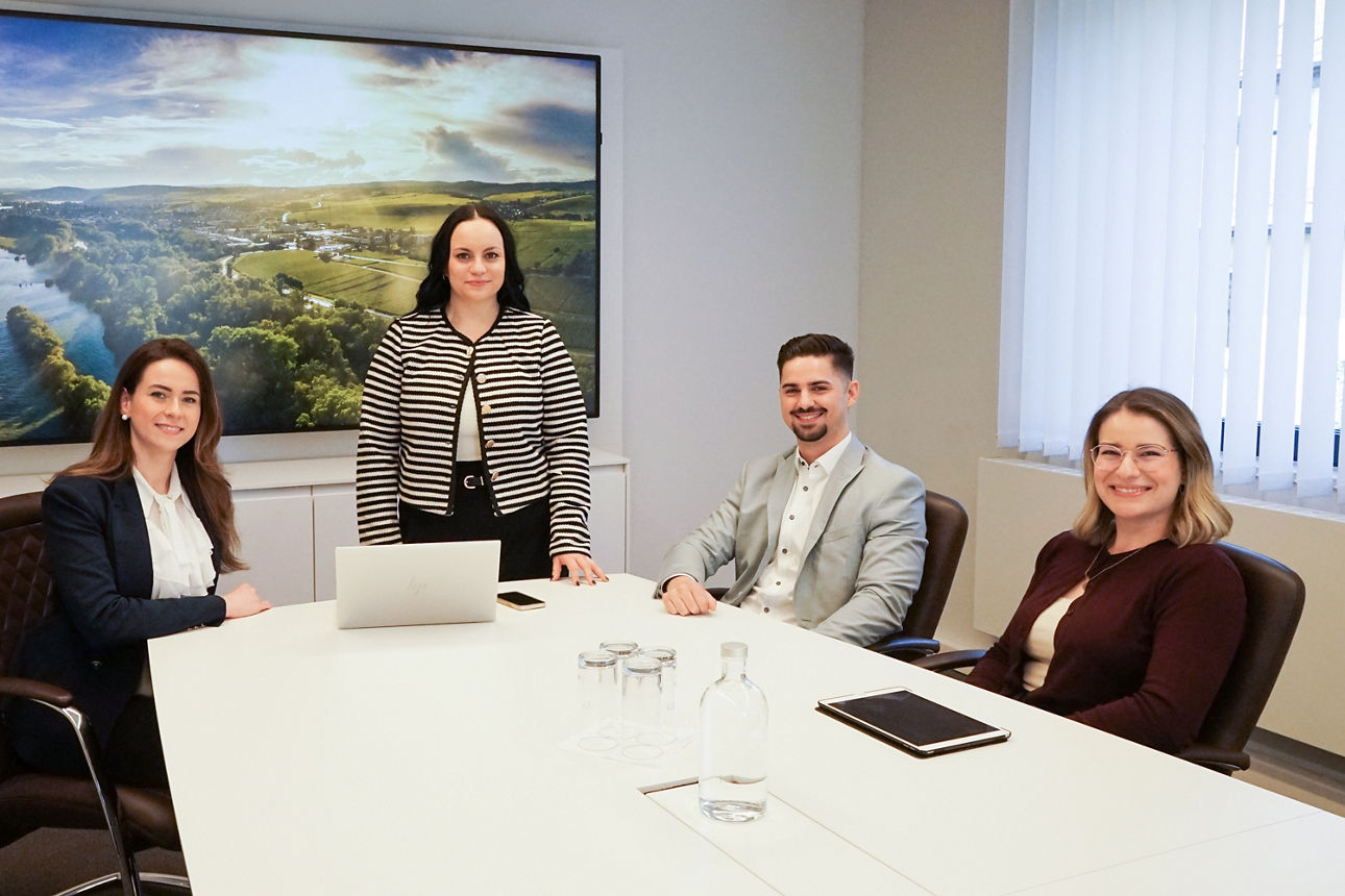  Vier Personen in einem Besprechungsraum, drei sitzen am Tisch, eine steht, alle lächeln, Laptop und Notizbuch auf dem Tisch                              