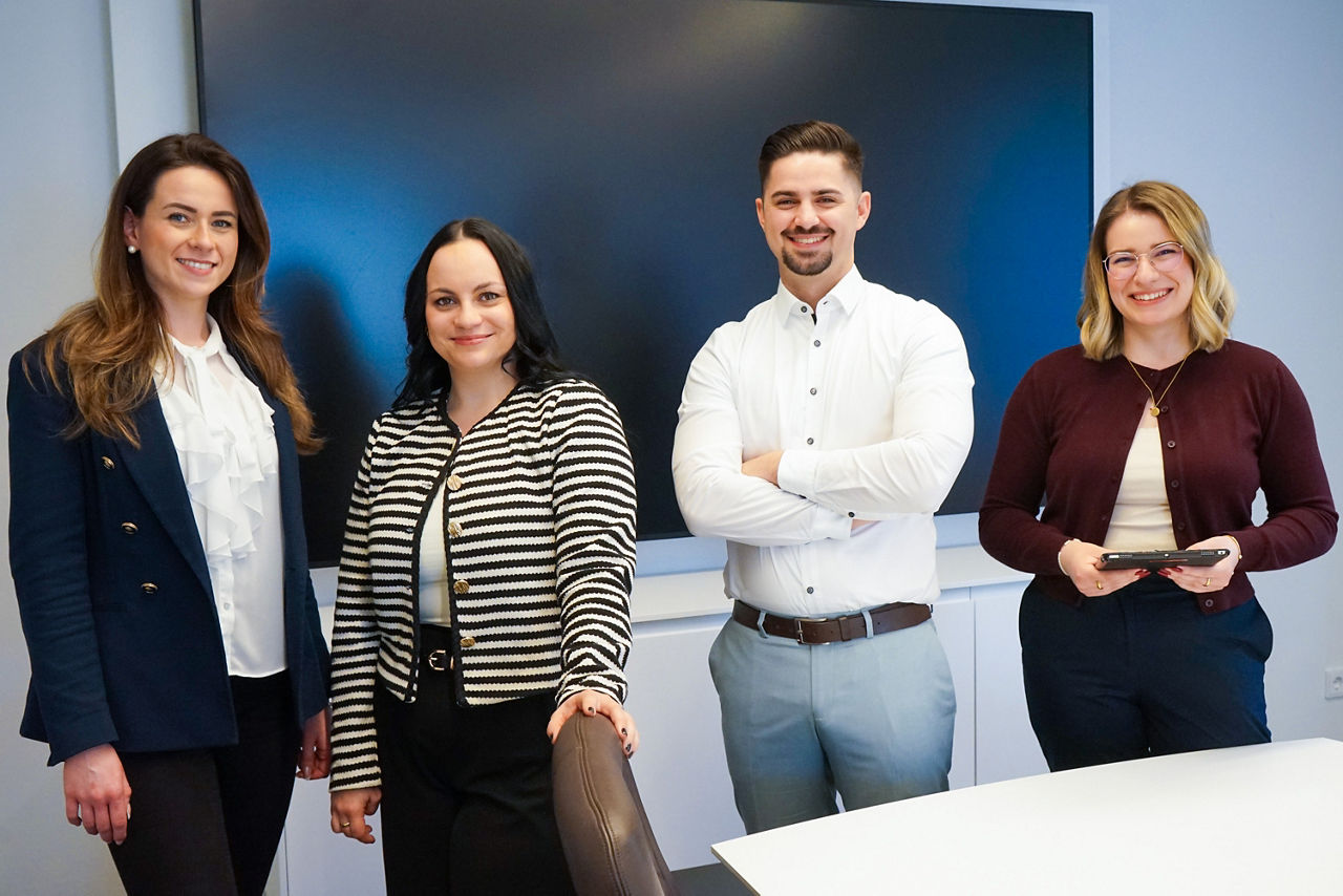 Vier junge Berufstätige, zwei Frauen links, ein Mann und eine Frau rechts, stehen in einem modernen Büro vor einem großen Bildschirm und lächeln in die Kamera.                               