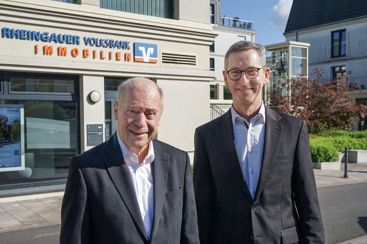 Zwei Männer in Anzügen stehen vor dem Gebäude der Rheingauer Volksbank Filiale in Eltville bei sonnigem Wetter
