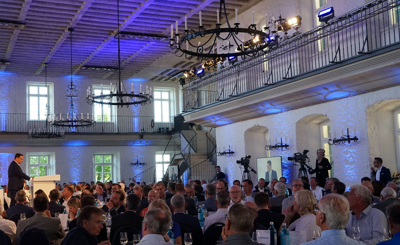 Großer Saal mit vielen Menschen an langen Tischen, Herr Zeiselmaier steht am Podium, Beleuchtung in Blau und Weiß