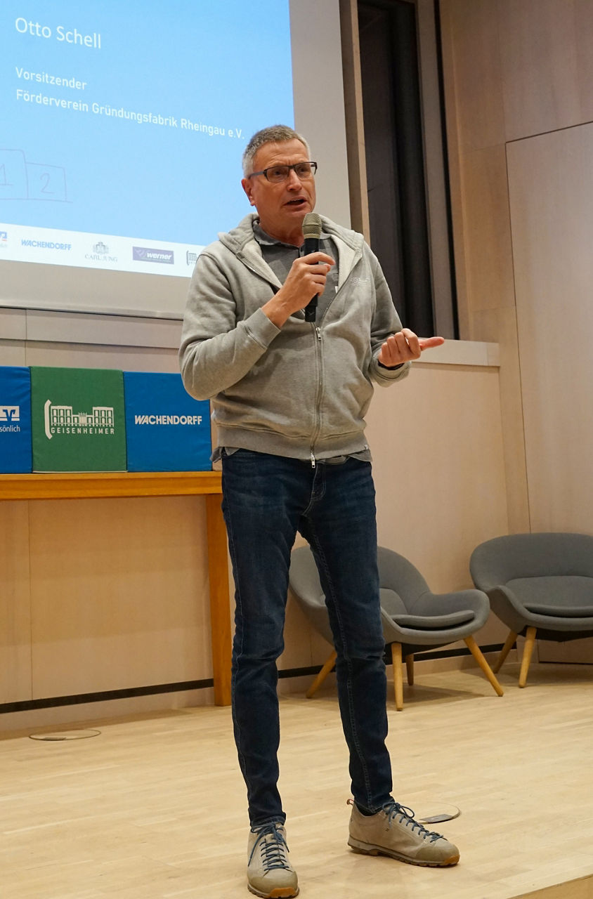 Mann in grauer Kapuzenjacke, blauer Jeanshose und grauen Schuhen hält Mikrofon in einem Vortragssaal mit Stühlen und Präsenta