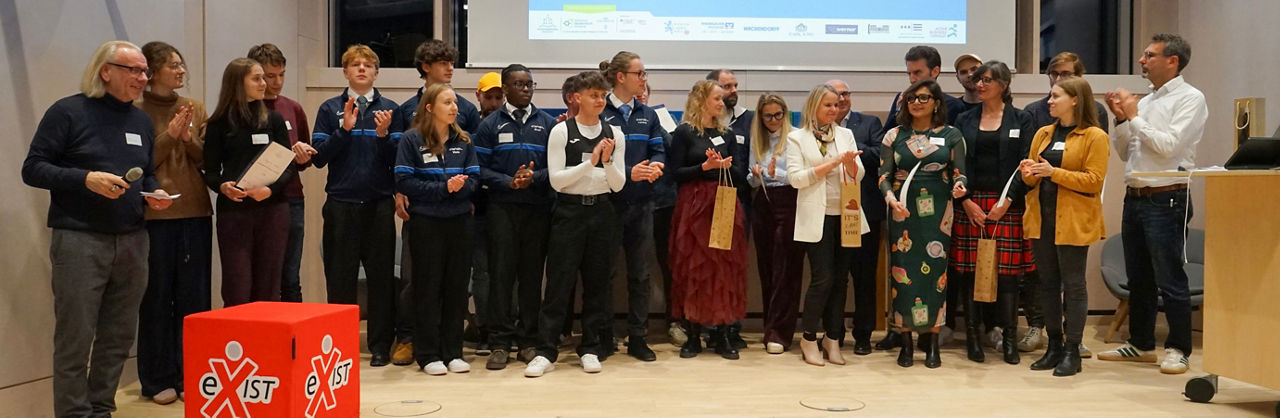 					Gruppe von Menschen steht nebeneinander auf einer Bühne, einige halten Urkunden