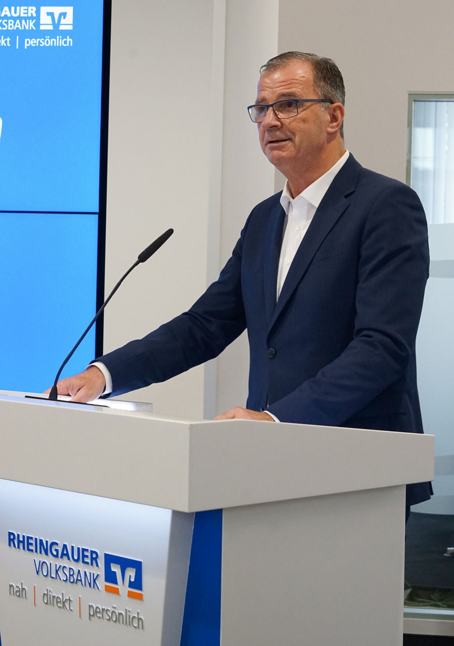 Mann in dunkelblauem Anzug und weißem Hemd steht an einem Rednerpult mit dem Logo der Rheingauer Volksbank. 
