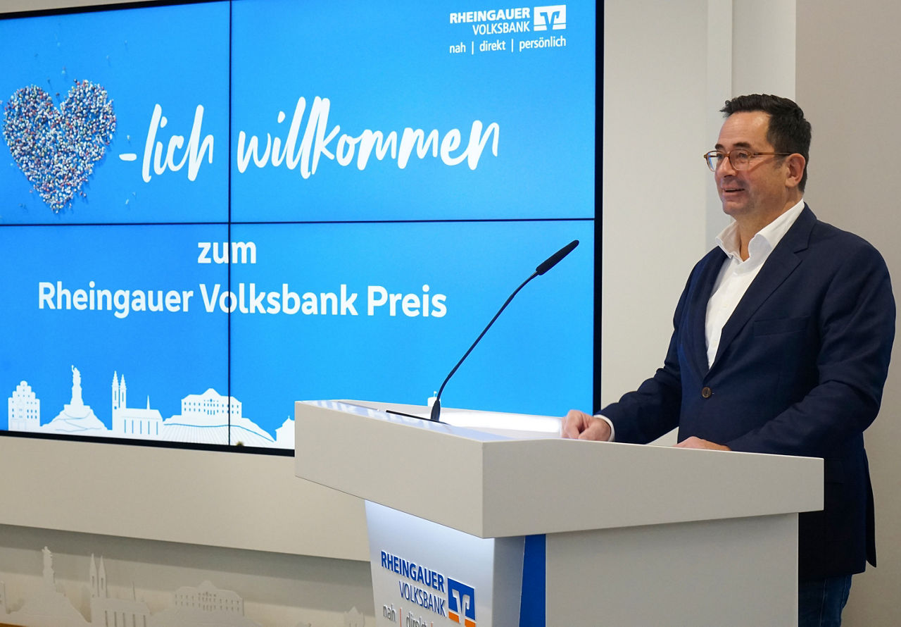 Mann im dunklen Sakko steht am Rednerpult mit Mikrofon vor großem Bildschirm mit Text 'Herzlich willkommen zum Rheingauer Volksbank Preis' 