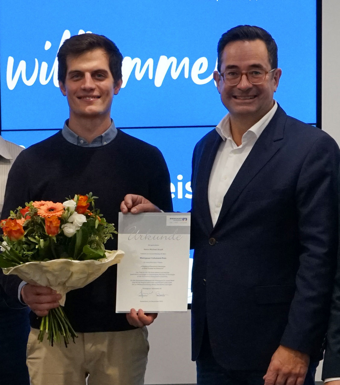 Zwei Männer stehen nebeneinander, einer hält einen Blumenstrauß, der andere eine Urkunde mit dem Wort 'Urkunde' oben.                               