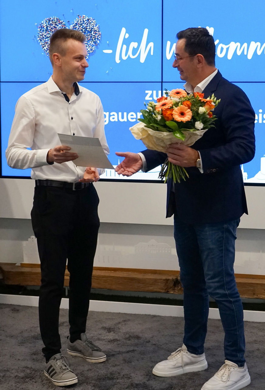 Zwei Männer stehen sich gegenüber, einer hält einen Blumenstrauß mit orangefarbenen und weißen Blumen, der andere ein Blatt Papier.                               