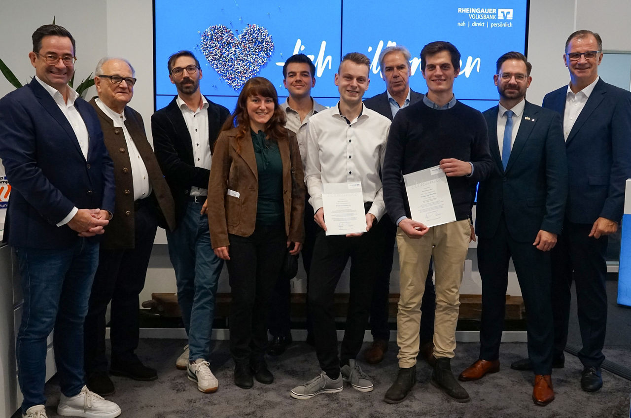 				Gruppe von zehn Personen, darunter zwei junge Männer in der Mitte mit Urkunden, vor blauem Bildschirm mit Rheingauer Volksbank Logo und Text.	