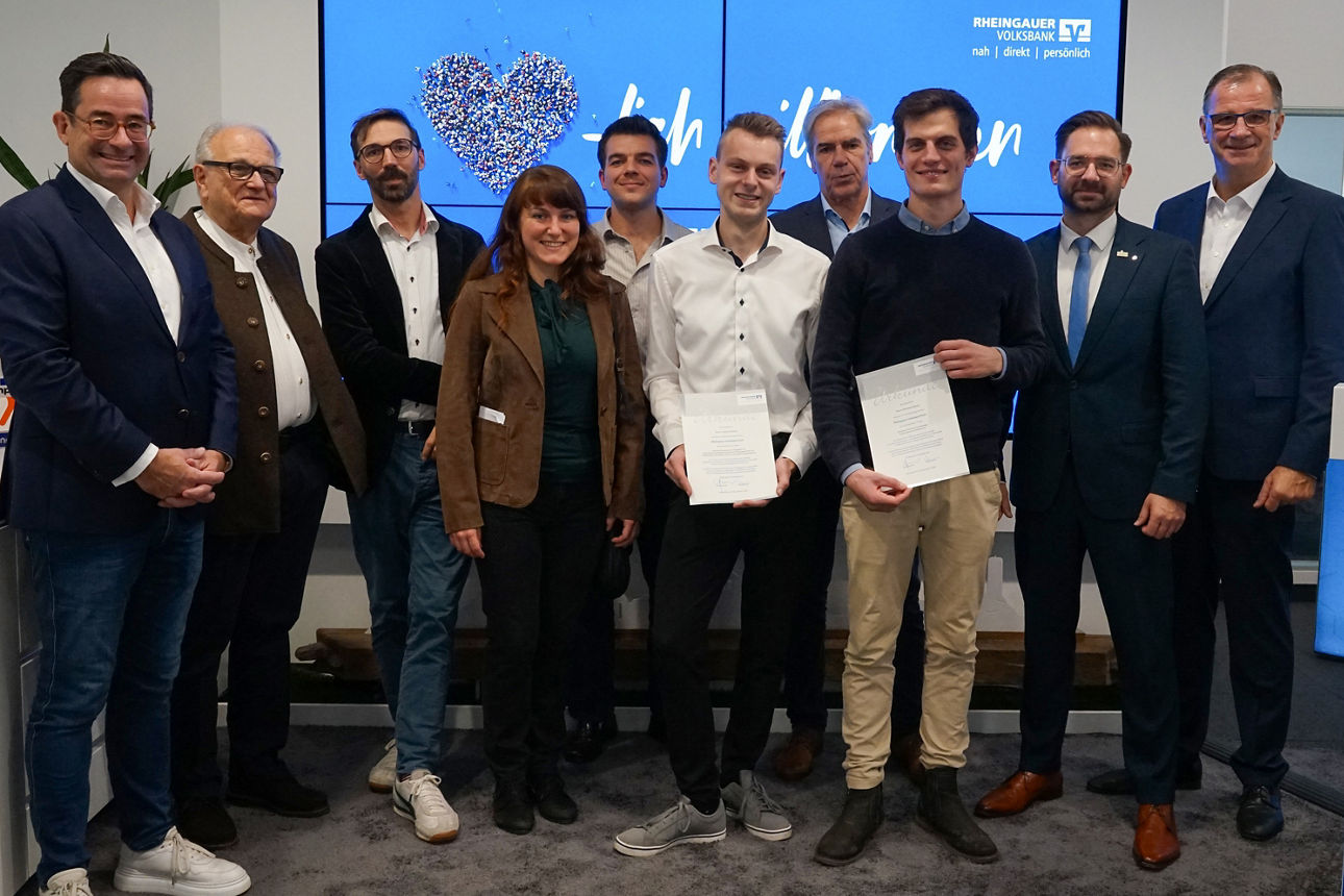 				Gruppe von zehn Personen, darunter zwei junge Männer in der Mitte mit Urkunden, vor blauem Bildschirm mit Rheingauer Volksbank Logo und Text.	