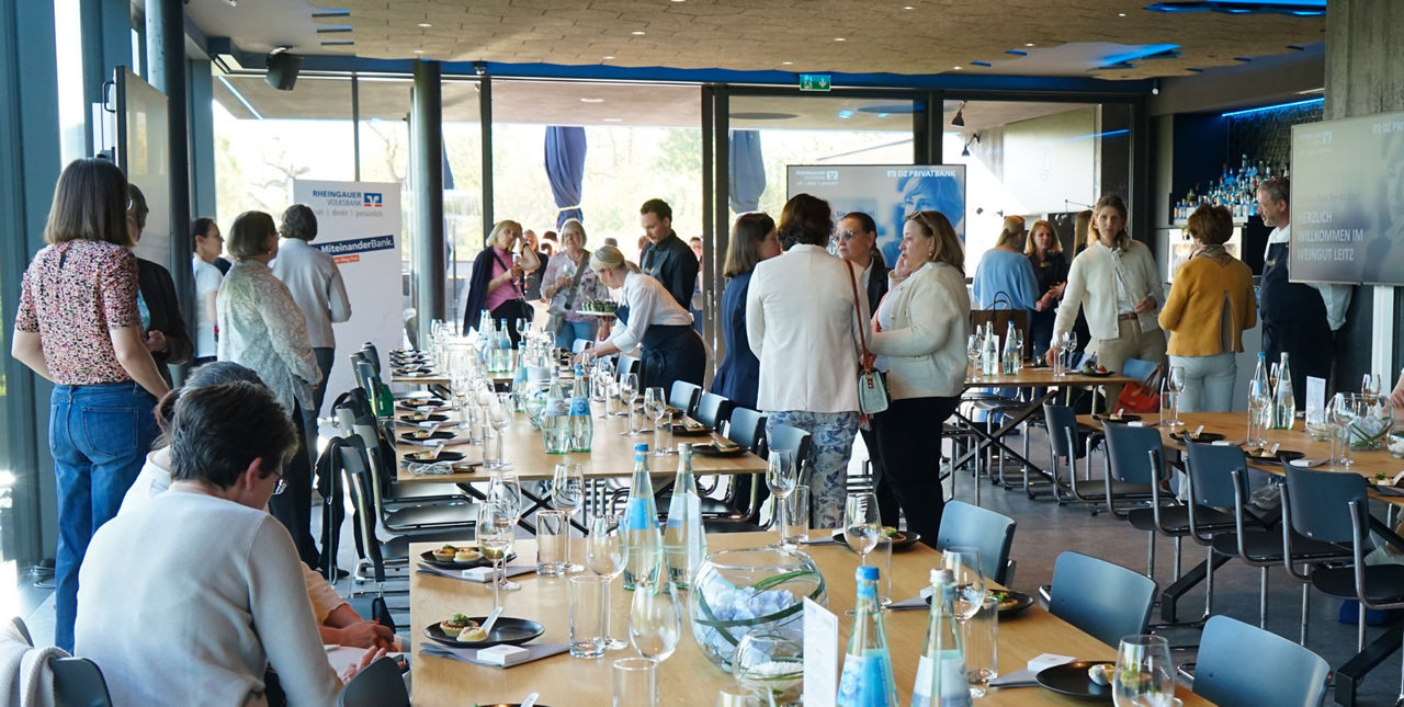 Menschen stehen und sitzen in einem hellen Raum mit großen Fenstern an einem langen Tisch, der mit Gläsern, Wasserflaschen und Tellern gedeckt ist                               