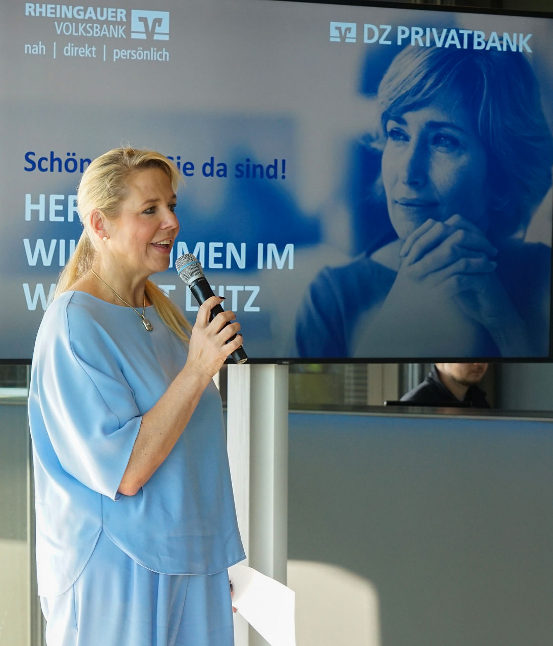  Frau in hellblauer Kleidung hält Mikrofon und Papier vor großem Bildschirm mit Text und Logos von Rheingauer Volksbank und DZ Privatbank                              